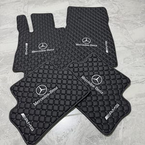 Puede incluir: Juego de alfombrillas negras para coche con diseño acolchado. Cada alfombrilla presenta el logotipo de Mercedes-Benz y el logotipo de AMG en blanco. El juego incluye alfombrillas para los asientos delanteros y traseros.