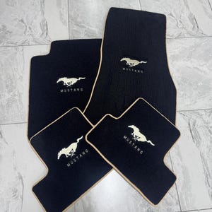 Può includere: Set di quattro tappetini neri per auto con bordo beige. Ogni tappetino presenta un logo Mustang bianco e la scritta "Mustang". I tappetini sono progettati per proteggere l'interno del veicolo.