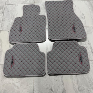May include: Set of four gray car floor mats with a quilted pattern and red stitching. Each mat has a white border and a "WORKS" logo in red and white. The mats are designed to fit a vehicle's interior.