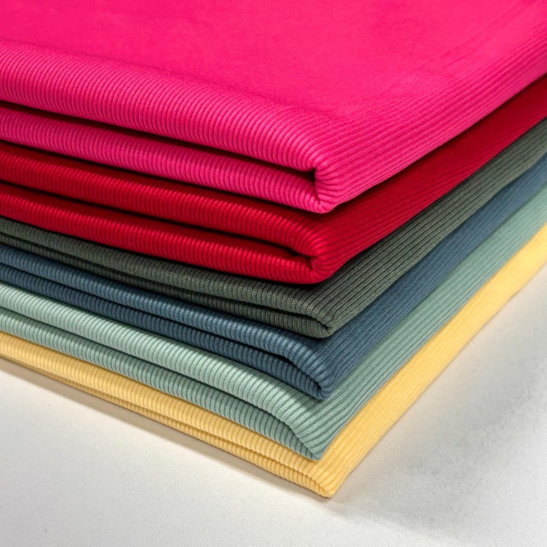 Op de afbeelding: Een stapel van zes verschillende geribbelde stoffen in verschillende kleuren. De stoffen zijn netjes op elkaar gevouwen, met de bovenste stof in een fel roze. De andere stoffen zijn rood, donkergroen, blauw, lichtblauw en geel.