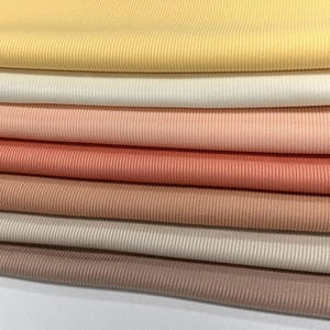 Op de afbeelding: Een stapel van zeven verschillende geribbelde stoffen. De kleuren zijn geel, wit, perzik, koraal, lichtbruin, beige en bruin.