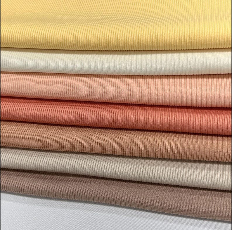 Op de afbeelding: Een stapel van zeven verschillende geribbelde stoffen. De kleuren zijn geel, wit, perzik, koraal, lichtbruin, beige en bruin.