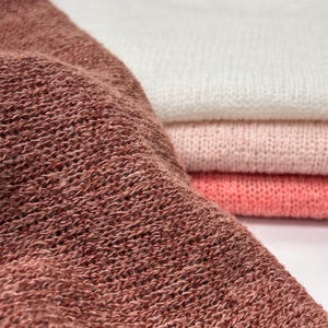 A close-up shot of a textured reddish-brown knitted fabric draped in the foreground with a neat stack of white, light pink, and coral knit fabrics blurred in the background.