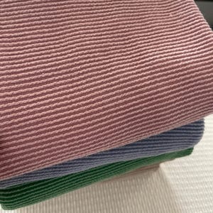 A close-up of three folded, ribbed knit fabrics in dusty rose, denim blue, and kelly green, stacked on a white textured surface.