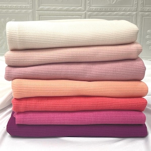 May include: A stack of waffle knit fabric in various shades of pink and orange. The fabric is folded neatly and arranged in a gradient from white to purple.