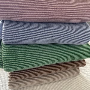 A stack of five folded, ribbed knit fabrics in dusty rose, denim blue, kelly green, coffee, and taupe, on a white textured surface.