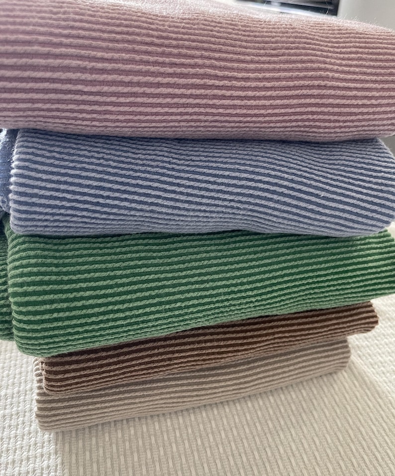 A stack of five folded, ribbed knit fabrics in dusty rose, denim blue, kelly green, coffee, and taupe, on a white textured surface.