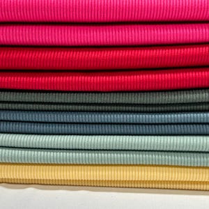 Op de afbeelding: Een stapel stofstalen in verschillende kleuren, waaronder roze, rood, grijs, blauw en geel. De stof heeft een geribbelde textuur.