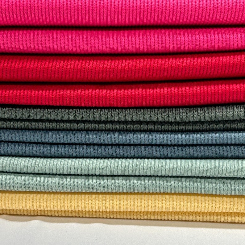Op de afbeelding: Een stapel stofstalen in verschillende kleuren, waaronder roze, rood, grijs, blauw en geel. De stof heeft een geribbelde textuur.
