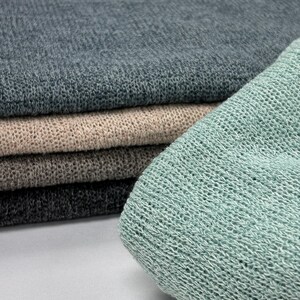 A folded mint-green knit fabric sits in the foreground with a stack of dark grey, beige, brown, and charcoal knitted fabrics resting behind it.