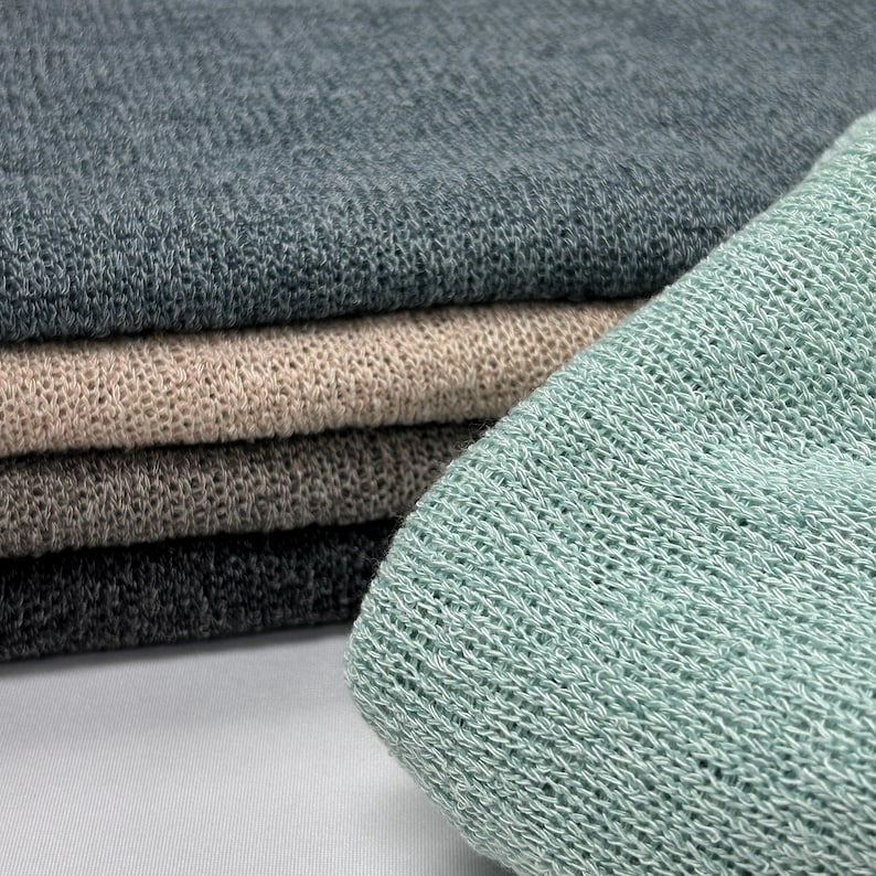 A folded mint-green knit fabric sits in the foreground with a stack of dark grey, beige, brown, and charcoal knitted fabrics resting behind it.