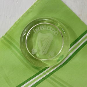 May include: A clear glass ashtray with the word "Ireland", a harp, and shamrocks etched into the glass. The ashtray sits on a green and white striped cloth.