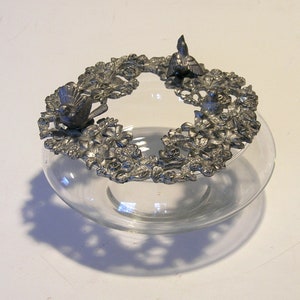 May include: A clear glass bowl with a decorative metal rim featuring three bird figurines and floral accents. The rim is silver-colored and encircles the bowl's opening. The bowl sits on a white surface.