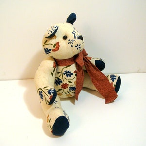 May include: A stuffed bear toy made from cream-colored fabric with a floral pattern in blue, red, and green. The bear features solid dark blue fabric on the ears, paws, and feet, a black button eye, and a long, solid rust-colored fabric scarf.