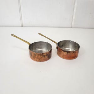 May include: Two small copper saucepans with brass handles. The pans have a round shape and are made of copper with a tarnished finish. The handles are long and thin, and the interior of the pans is a shiny silver color. The saucepans are sitting on a white surface.