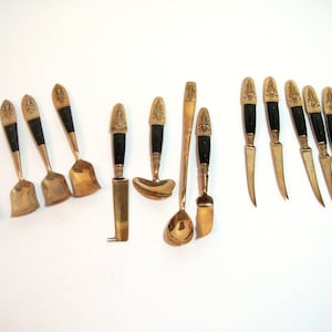 May include: A collection of antique serving utensils, including spoons, knives, and spreaders. Each piece features a brass-colored blade or bowl, a black handle, and a decorative gold-colored figure at the top. The utensils are arranged on a white surface.