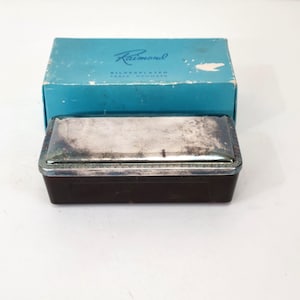 May include: A silver-plated table crumber with a rectangular shape and a dark brown base. The lid is silver with a decorative border. The box is turquoise with the name "Raimond" printed on it.