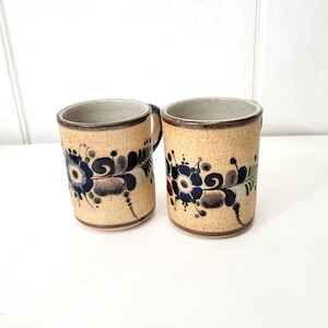 May include: Two cylindrical ceramic mugs with a speckled beige base and a dark brown rim. Each mug features a hand-painted floral design in shades of dark blue, black, and muted green, with swirling leaf patterns encircling the exterior surface.