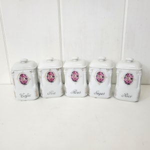 May include: Five white ceramic kitchen canisters with lids, labeled "Coffee", "Tea", "Flour", "Sugar", and "Rice". Each canister features a floral design with pink and purple roses and gold accents.