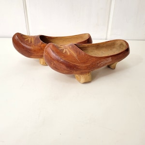 May include: A pair of wooden clogs with carved floral designs. The clogs are brown and have a rounded toe. They are sitting on a white surface.