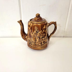 May include: A brown and cream ceramic teapot with a raised design of a woman drawing water from a well. The teapot has a brown glaze with a mottled effect and the text "Drink all this wine" is visible on the base.
