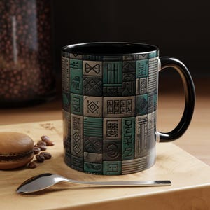 May include: A ceramic coffee mug with a black handle and interior. The mug features a repeating pattern of teal, gray, and black geometric shapes. The mug sits on a wooden surface with a spoon and a pastry.