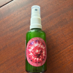 May include: A green glass bottle with a white spray top and a label that says "The Rose Alchemist Diamond Eye of Queen of Hearts". The label features a red rose with a sparkling background.