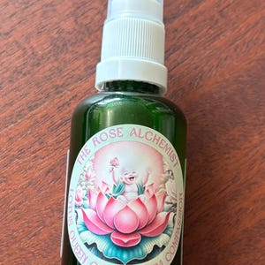 Op de afbeelding: Groene glazen spuitfles met een witte dop. Het etiket toont een lotusbloem ontwerp met de tekst "The Rose Alchemist, Queen of Quantum Compassion."