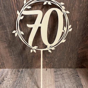 May include: A wooden cake topper featuring the number 70 in a stylized font, encircled by a wreath of leaves. The topper is light beige and has a long, pointed stake for insertion into a cake. The design is suitable for a 70th birthday celebration.
