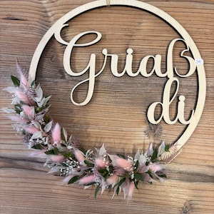 May include: A wooden wreath with the words "Grials di" in cursive script. The wreath is decorated with dried pink and white flowers.
