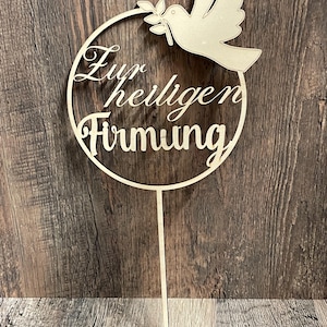 Puede incluir: Adorno de pastel de madera con el texto alemán "Zur heiligen Firmung" dentro de un círculo. Una paloma que lleva una rama de olivo está en la parte superior. El adorno está hecho de madera clara y tiene un palo puntiagudo para insertar.