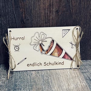 May include: A light-colored wooden plaque with the words "Hurra!" and "endlich Schulkind" engraved on it. The plaque features a laser-cut design of a school cone filled with money, a clock, a pencil, a triangle, and scissors. The plaque is tied with twine.