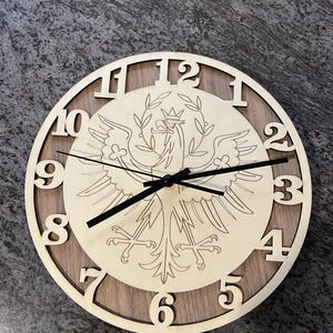 May include: A wooden clock with a brown and light wood finish. The clock face features a white eagle with a crown and a wreath of leaves. The numbers 1 through 12 are around the edge of the clock face.