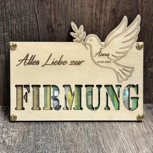 Puede incluir: Una placa de madera con las palabras "Alles Liebe zur FIRMUNG" recortadas. Una paloma con una rama de olivo está en la parte superior derecha. El nombre "Anna" y la fecha "24.05.2025" también están en la placa. Las letras de la palabra "FIRMUNG" están llenas de dinero colorido.
