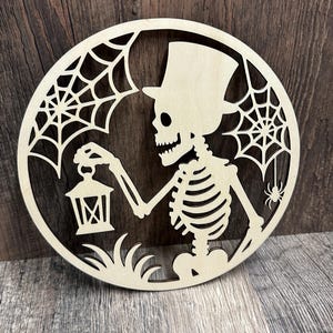 May include: A laser-cut wooden Halloween decoration featuring a skeleton wearing a top hat and holding a lantern. The skeleton is surrounded by a spider web design and grass. A small spider is also present. The decoration is circular.