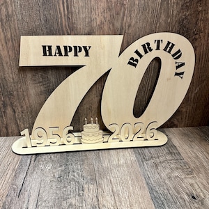 May include: Wooden birthday sign with the words "HAPPY BIRTHDAY 70" cut out. The number 70 is large and central, with the years 1956 and 2026 below, and a small cake with candles. The sign is made of light-colored wood.