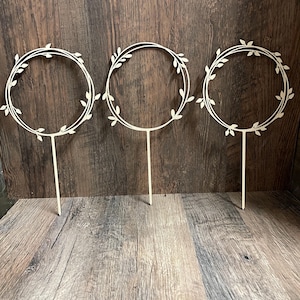 Puede incluir: Tres adornos de pastel de madera con un diseño de corona circular. Cada corona está adornada con delicados detalles de hojas y montada sobre un fino palito. Los adornos son de color claro y están colocados sobre un fondo de madera.