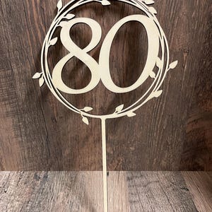 Könnte beinhalten: Ein Kuchenaufsatz aus Holz mit der Zahl 80 in dekorativer Schrift, umgeben von einem Blätterkranz. Der Aufsatz hat einen langen, dünnen Stab zum Einstecken in einen Kuchen. Das Holz hat eine natürliche, hellbraune Farbe.