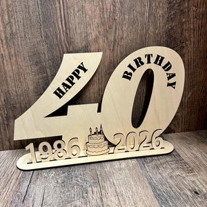 Op de afbeelding: Een houten verjaardagsdecoratie met het getal 40, de woorden "HAPPY BIRTHDAY" en de jaren 1986 en 2026. Een kleine taart met kaarsen is ook inbegrepen. De decoratie is gemaakt van lichtgekleurd hout.
