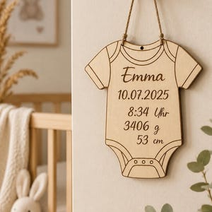 Pode incluir: Um enfeite de parede de madeira clara em forma de body de bebé, suspenso por um cordão fino. A madeira está gravada com o nome Emma, a data 10.07.2025, a hora 8:34 Uhr, o peso 3406 g e o comprimento 53 cm em letras castanho-escuras.