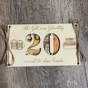 May include: A wooden birthday card with the number "20" cut out, revealing currency. The card features the text "Alles Gute zum Geburtstag" and "w&uuml;nscht dir deine Familie." Decorative elements include beer mugs and a cake with candles.