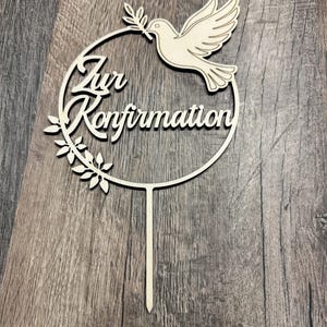 Può includere: Un cake topper in legno con la scritta "Zur Konfirmation". Il topper presenta una colomba con un ramo d'ulivo, all'interno di una cornice circolare con foglie. Realizzato in legno chiaro.
