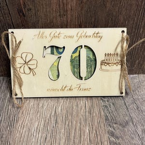 以下が含まれることがあります： 70の数字がくり抜かれた木製の誕生日カード。カードには、四つ葉のクローバー、ろうそくのケーキ、テキスト「Alles Gute zum Geburtstag wünscht die Franz」が描かれています。ジュートの紐が取り付けられています。