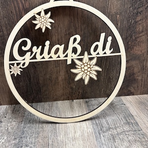 May include: A circular light-coloured wooden wall hanging featuring the laser-cut text "Griaß di" across the centre. The design includes three detailed edelweiss flower cutouts positioned around the text within the circular wooden frame.