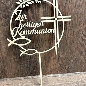 May include: A wooden cake topper with the German text "Zur heiligen Kommunion" (For Holy Communion). The design includes a cross, two fish, and a floral wreath, all in a light wood color.