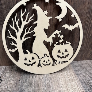 May include: A wooden Halloween decoration featuring a witch stirring a cauldron, with pumpkins, bats, a tree, stars, and a crescent moon. The circular design is cut from light-colored wood.