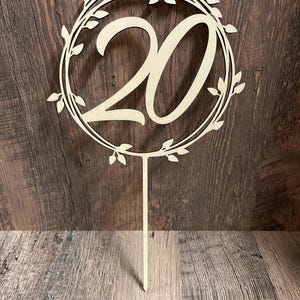 Peut inclure: Un cake topper en bois clair représentant le chiffre 20 dans une police décorative, entouré d'une couronne de feuilles. Le topper est en bois clair et possède une longue tige pointue pour l'insérer dans un gâteau.