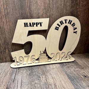 以下が含まれることがあります： 木製の誕生日飾りで、数字の50、"HAPPY BIRTHDAY"の文字、1976年と2026年が書かれています。年の間に、ろうそくのついた小さなケーキがあります。テキストは黒色です。