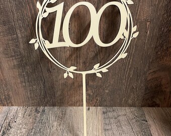 May include: A wooden cake topper with the number "100" in a decorative font, encircled by a wreath of leaves. The topper has a long, thin stake for insertion into a cake. The design is in a natural wood tone.