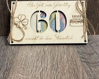 May include: A wooden birthday card with the number 60 cut out, revealing colorful paper beneath. The card has the text "Alles Gute zum Geburtstag" and "wünscht dir dein Stammtisch" engraved on it, along with a four-leaf clover and a cake with candles.